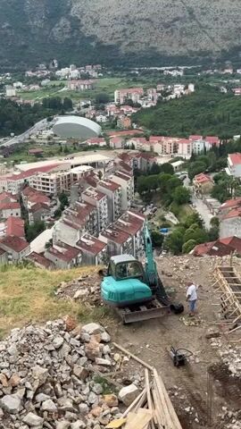 A wide shot shows a construction site on a hillside overlooking a town. In the foreground, a turquoise excavator is parked on a dirt surface littered with rocks and wooden planks. A person wearing a white shirt and blue shorts stands next to the excavator, appearing to be working on it. A plate compactor is visible in the background. In the mid-ground, the excavator is positioned on a slope with a partially constructed wall made of wood forms and rebar. Beyond the construction site, a town with multi-story buildings with red roofs stretches out, nestled against a lush green hillside. A large, white, dome-shaped structure is visible in the distance. The sky is overcast.