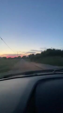 The video is filmed from inside a vehicle, looking forward through the windshield. The view is of a rural road at dusk. The sky is a gradient of blue to pale orange and pink near the horizon. There are telephone poles and wires visible along the side of the road. Fields and trees line the road, with some scattered houses or buildings in the distance. The road appears to be paved and is wet in places, suggesting recent rain. The dashboard of the vehicle is visible in the foreground. The camera is stationary, implying the vehicle is moving forward. There are no people visible in the frame.