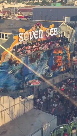 This is a high-angle video of a fairground ride called "Seven Seas". The ride is a swinging boat that is currently in motion, with passengers seated and facing outwards. The backdrop of the ride is a colorful mural depicting a pirate ship in stormy seas. Below the ride, a crowd of people mills around, some watching the ride, others walking through the fair. The atmosphere is lively and bustling. The sun is shining, casting long shadows, and the overall scene suggests a typical summer day at a fair.