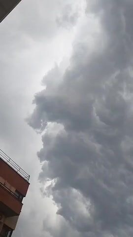 The video shows a close-up, vertical view of a dramatic, overcast sky filled with dark, turbulent storm clouds. The clouds are dense and textured, with swirling patterns and varying shades of gray, indicating an impending storm or a recent weather event. The lighting is diffused and somber, suggesting it is either early morning or late afternoon under heavy cloud cover.

In the bottom left corner of the first frame, a sliver of an apartment building is visible, with a balcony railing and an orange-toned facade. This provides a subtle hint of an urban environment. The rest of the footage focuses entirely on the sky and clouds, creating a sense of awe and perhaps a touch of foreboding due to the intensity of the weather. There is no visible text in the video. The overall mood is one of natural power and the anticipation of inclement weather.