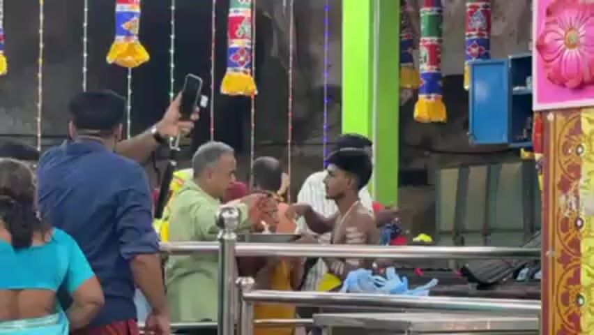 The video depicts a group of people inside what appears to be a temple. Most are standing behind a metal railing. Several individuals have their hands together in a prayerful position. A younger man stands shirtless with white markings on his body. An older man in a light green shirt touches the young man's head. Another man in a blue shirt holds up his phone, possibly taking a photo or video. Colorful decorations, including garlands, hang from the ceiling. A blue cabinet is visible in the background. There is a general air of reverence and religious activity.
