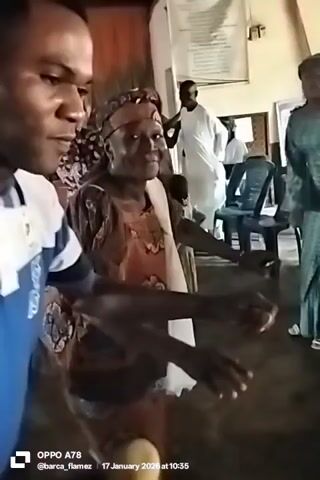 The video shows several people in a room, perhaps at an event.  One man is filming the scene, focusing on an older woman with a headwrap and traditional garb, as they both dance in place to an unseen sound.  Other individuals, dressed in various styles including traditional white attire and a green outfit, are visible in the background, some seated. The room has a basic interior, with chairs and what looks like an informational poster on the wall.  The video is taken with a mobile phone that imprints the date "17 January 2026" and the time "10:35" on the video.