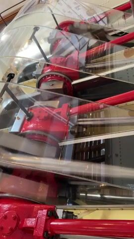 The video shows a close-up of a large, red industrial machine with metallic parts. The machine appears to be in motion, with various components rotating and moving. It has a complex structure with visible gears, pipes, and cylinders. The material is shiny and reflects light, suggesting it is made of metal. The background is blurred, but appears to be an indoor environment, possibly a museum or factory, with some wooden elements and more machinery visible. The lighting is bright and seems to come from above. The overall impression is that of a powerful and intricate piece of machinery in operation.