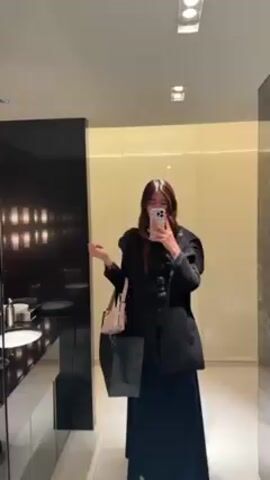 The video shows a person in a bathroom taking a selfie in a mirror. The person is wearing a black outfit with a jacket and a long skirt or dress and carrying a light-colored handbag and a black shopping bag.  The bathroom has a dark marble wall, a sink, and decorative sconces with white cylindrical shades and gold accents. The mirror is rectangular with a gold frame. The lighting in the bathroom is bright. The person is holding a smartphone and looking at the screen while taking the selfie. The bathroom has a modern and luxurious aesthetic.