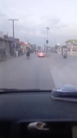 The view from inside a car traveling down a street in a town is presented. A silver car is ahead, followed by a couple of motorized rickshaws, and a motorcycle pulls out from the left. People are visible walking on the sidewalks. The street is paved and mostly deserted. Buildings line the road, and utility poles and streetlights are visible. The sky is overcast. The dashboard of the car from which the video is taken is dark and cluttered with papers. An alarm siren can be heard in the background.
