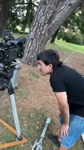 The video shows a man in his 20s, wearing glasses and a black shirt with short denim shorts, standing next to a professional film camera mounted on a tripod in a grassy area, possibly a park. The camera is large and complex with various attachments and screens. The man appears to be inspecting or adjusting the camera. There's a large tree in the background and an equipment case nearby. The lighting suggests it might be late afternoon or early evening. The scene appears to be a film set or a location for professional video shooting.