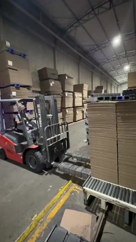 In a large warehouse with a high ceiling and a gray metal roof, a red and black HELI forklift is shown in action. The forklift is being operated by a person wearing a light-colored shirt and dark pants, who is seated and focused on the task at hand. The forklift's forks are elevated, holding a large stack of brown cardboard sheets that are being lifted from a white conveyor belt with metal rollers. The warehouse floor is gray, marked with yellow safety lines. To the left, stacks of flattened cardboard boxes and wooden pallets are stored. In the distance, more stacks of cardboard and blue plastic pallets are visible under the fluorescent lighting. The camera remains static, providing a clear view of the forklift operation.