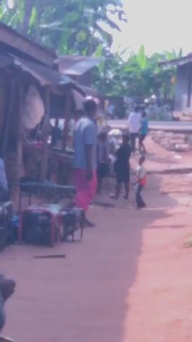 The video shows a street scene in what appears to be a village or small town. The camera follows a person pushing a wheelbarrow. In the background, there are small, rudimentary buildings or stalls. Several people are visible, including one wearing pink pants and a striped shirt. A generator is also visible near the camera's subject.
