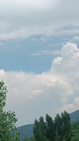 The video shows a wide shot of a cloudy sky and green hills. There are trees in the foreground and middle ground. The clouds are white and puffy, with some darker gray clouds in the distance. The sky is a light blue. The hills are covered in green trees.