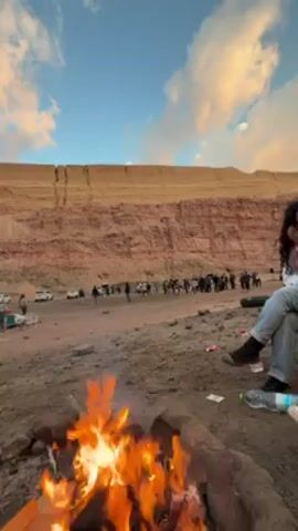 The video shows a campfire burning in a dry, sandy area. In the background, a large group of people are gathered near a line of parked cars at the base of a cliff. A person is sitting in a chair on the right side of the frame, near the campfire. The sky is blue with some clouds. There are some plastic water bottles on the ground near the campfire.