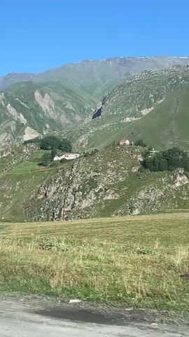 The video shows a scenic view of a mountain village. The foreground features a grassy field with some colorful objects, possibly beehives. The middle ground shows a cluster of houses and buildings nestled on a rocky, green hillside. In the background, a large, majestic mountain dominates the landscape, its slopes covered in lush greenery. The sky is clear and blue. The camera appears to be moving, suggesting the video was filmed from a vehicle.