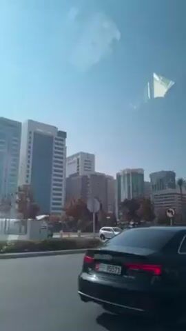The video shows a view of a city skyline through a car window.  Several tall buildings of varying heights and designs are visible, along with trees lining the street. Cars are driving on the road, and traffic signals are present. The sky is clear and blue. The car window has reflections on it.