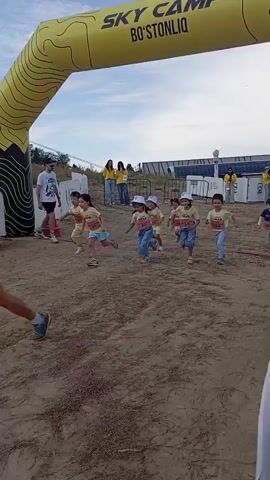 At timestamp 00:00, a group of young children wearing light-colored shirts and hats are running through an inflatable archway. The ground is dirt and sand. In the background, there are adult spectators and what appears to be a camera crew. The archway has yellow and black markings.

At timestamp 00:01, the children are continuing to run. They are now in a line, with some children ahead of others. There are white barriers with logos in the background.

At timestamp 00:02, the children are still running, and the terrain has become more uneven with dirt and rocks. An adult is running behind the children. There are orange and white traffic cones placed on the ground.

At timestamp 00:03, the children are spread out further, with some children at the front and others further behind. The adult is still in pursuit. The traffic cones are visible on the side of the path. The landscape includes hills covered with trees in the background.

At timestamp 00:04, the children are running up a slight incline. The group is more spread out now, and one child in a blue shirt is further ahead. The hills in the background are more prominent, with some buildings or structures visible on them. The sky is partly cloudy.