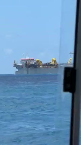 The video presents a view from inside a watercraft, likely a ship or a boat, looking out towards the ocean. Through a window pane, a large vessel, presumably a ship, is visible in the distance on the water. The window has a partially obscured view due to water droplets or residue on its surface. The sky is clear and blue. The ocean appears calm with minor ripples. The large vessel appears to be stationary in the water.