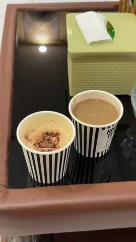 Two paper cups with drinks are placed on a dark, reflective surface, likely a table or tray. The surface has a brown, leather-like border visible on the left. In the background, a light green, textured tissue box is present.

The cup on the left contains a light brown, foamy drink topped with a generous sprinkle of dark powder, possibly cocoa or cinnamon, forming a roughly heart-shaped pattern. This suggests a coffee beverage like a cappuccino or latte. The cup on the right holds a uniform, medium brown liquid, which could be tea with milk or another coffee drink. Some text is visible on the right cup, starting with "HIG", likely part of "HIGH TEA" or similar branding.

Both cups feature a distinctive black and white vertical stripe design. The reflective surface clearly shows reflections of the cups and a bright, circular light source from above. The setting appears to be indoors, possibly in a cafe or lounge, with artificial lighting. No people are visible, and no specific activity beyond the presentation of the drinks is shown.