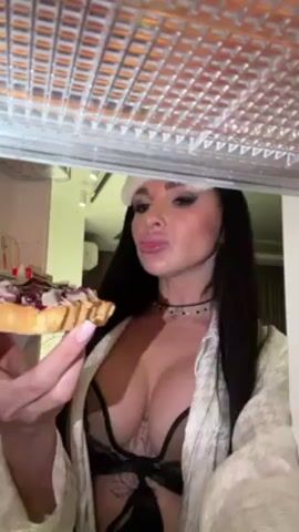 A woman with long, dark hair, wearing a lacy bra, white robe, and a white eye mask on her forehead, stands in front of an open refrigerator. She's holding a small tart or pastry in her hands and takes a bite of it. The inside of the refrigerator is visible, with various items on the shelves.