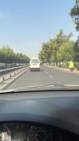 The video shows a POV shot from inside a vehicle traveling on a road. The camera is looking forward through the windshield. In the distance, a white van is driving in the same direction. The road is paved and appears to be a multi-lane highway with a guardrail on the left side. Tall green trees line the right side of the road, and the sky is clear and blue. The interior of the vehicle is visible in the foreground, including the dashboard and part of the steering wheel. The video appears to be a continuous shot with no visible edits or camera movements other than what would occur from driving.