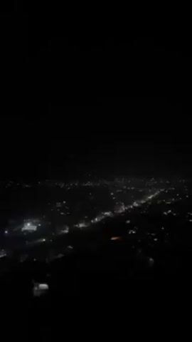 The video shows a nighttime view of a town from a high vantage point. The sky is dark, and the town below is illuminated by streetlights and building lights. Some trees are visible in the foreground. The camera is fairly still, offering a static view of the landscape. There is no clear activity taking place in the frame.
