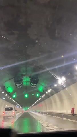 The video shows the interior of a tunnel, viewed from the perspective of a vehicle driving through it. The tunnel is well-lit with rows of white lights along the sides and green lights illuminated along the ceiling, indicating a clear path forward. Two large metallic cylindrical objects are visible hanging from the ceiling at the beginning of the video, possibly part of the tunnel's ventilation system. The road surface appears wet, suggesting it might be raining or has recently rained, and there are visible water droplets on the windshield. A white van is traveling ahead in the right lane. Green arrows are displayed on the ceiling, pointing forward, and the tunnel appears to be a multi-lane highway. The video lasts for approximately five seconds.