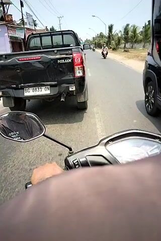A first-person perspective video shows a person riding a scooter on a paved road. In front of the scooter is a black pickup truck with the license plate BG 8831 ON. To the right of the pickup truck, on the same road, are several other motorcycles and a white car. The road appears to be in a rural or suburban area with palm trees visible on the right side of the road. The sky is clear and the sun is shining. The person on the scooter is wearing a helmet and a dark-colored shirt. Their hand is visible on the scooter's handlebar.