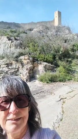 The video shows a woman taking a selfie. The woman is wearing sunglasses and a light pink sweater. She has gray hair. Behind her is a rocky hillside with sparse vegetation. At the top of the hill is a stone tower. The sky is clear and blue. The woman is smiling. The video appears to be taken outdoors on a sunny day.