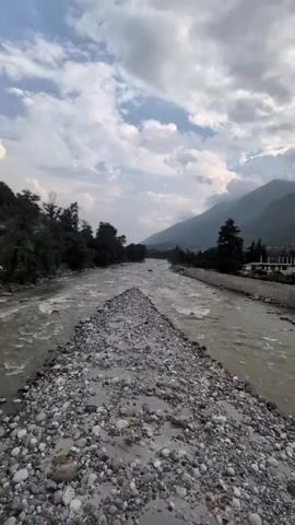 The video shows a motorcyclist crossing a bridge over a river. The river is wide and shallow, with a rocky bottom. The banks of the river are lined with trees and mountains. The sky is cloudy. The motorcycle is a Royal Enfield, and the rider is wearing a helmet. The bridge has black and white striped railings. The river is a light brown color, and the water is moving quickly. There are some buildings visible on the far side of the river, in the mountains.