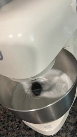 A white stand mixer is in operation. The mixing attachment, a whisk, is churning a white, frothy substance in a stainless steel bowl. The mixture is creating a vortex as it is whipped. The stand mixer is white with a silver trim around the bowl. The background shows a speckled countertop.
