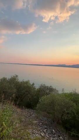 This is a video of a sunset over a large body of water, likely a lake. The scene features the calm water reflecting the warm hues of the sunset, with a gentle shoreline dotted with trees and what appears to be a small beach with loungers. In the foreground, there is lush green foliage and rocky terrain. The sky is painted with a gradient of soft pinks, oranges, and blues, with scattered clouds catching the last rays of sunlight. Distant mountains form a silhouette against the horizon. The overall atmosphere is peaceful and serene, characteristic of a beautiful evening in a scenic location. The time of day is clearly sunset. The weather appears to be calm and clear, with gentle cloud formations. The location cues, like the type of vegetation and the appearance of the mountains and lake, are consistent with a Mediterranean setting, specifically Garda, Italy, as indicated by the metadata. There is no visible text in the video.