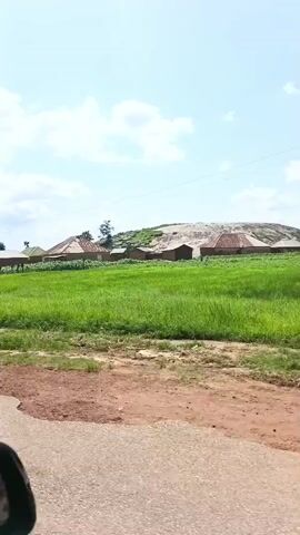 The video shows a car driving on a dirt road through a rural area. On the left side of the road, there are green fields, some scattered buildings with corrugated metal roofs, and a large rocky outcrop in the distance. The sky is blue with scattered white clouds. On the right side of the road, there are more green fields and some buildings. The camera appears to be mounted inside the vehicle, possibly on the dashboard or windshield, as the side mirror is visible in the lower left corner of the frame. The overall impression is of a developing or undeveloped area with simple housing and abundant vegetation.