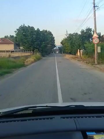 The video is a first-person perspective from inside a vehicle, showing a road lined with trees and some buildings. The road has a white dividing line. The sky appears clear and bright, suggesting daytime. There are power lines visible along the side of the road. The vehicle appears to be moving forward at a moderate speed. The scene is consistent with driving through a rural or suburban area. No people are visible within the frame.