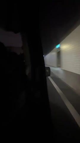From inside a moving vehicle, the perspective looks out the passenger-side window. The vehicle exits a brightly lit tunnel with white tiled walls. Outside the tunnel, the sky is a dusky blue, and streetlights are visible. A long, modern building runs parallel to the road. The white lines of the road lane markings are visible. Inside the vehicle, a person is silhouetted against the window, wearing a dark jacket.