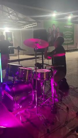 A person is playing a drum set under pink and green lights. A speaker is to the right of the drum set and a person is standing near the speaker. The background shows a banner with text in Russian and images of people. The scene appears to be outdoors at night.