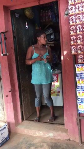 A woman is standing in the doorway of a small store. She has dark hair styled in small braids and is wearing a light blue tank top, cropped gray leggings, and flip-flops. She holds a small bill of currency in her right hand, extending it slightly toward the camera. Behind her, the store's interior is visible, packed with various items including drinks, packaged goods, and a small refrigerator. The store's entrance is framed by a red doorway. A display rack stocked with neatly arranged packets of goods is visible on the right. The floor of the doorway appears to be concrete. A box is visible on the ground to the left.