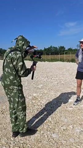 A man in camouflage records a man and a woman in athletic attire standing in front of a white banner with colorful logos. The man recording is positioned with his back to the viewer, holding a camera on a gimbal. The man and woman are smiling and posing for the camera on a gravel-covered ground under a clear blue sky.