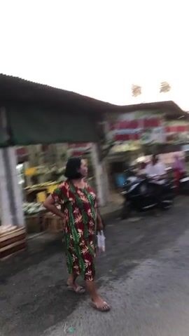 The video shows an outdoor scene, likely a street or market area, with several buildings and parked motorcycles visible. The lighting suggests it is either early morning or late afternoon.

In the foreground, a woman wearing a patterned dress is walking across the street. She is facing to the left of the frame. Her right hand is on her back. Her left arm is extended slightly forward.

To the left of the woman, there is a building with a dark roof and an open front. Signs are visible on this building, one of which appears to read "ES SELAMAT MOED". There are also displays of produce, possibly fruits or vegetables, on tables outside this building. A woman in a dark outfit is standing near the produce. A motorcycle is parked in front of this building.

Further down the street, more buildings and parked motorcycles can be seen. There are power lines running overhead. In the background, trees and vegetation are visible. The sky is overcast with some lighter areas suggesting the direction of the sun. Two motorcycles are moving away from the camera on the right side of the street.
