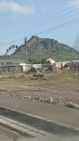 The video shows a daytime outdoor scene with a large rocky mountain in the background. In the foreground, there is a dirt lot with piles of gravel and construction materials. Several low-rise buildings and fences are visible, along with some vegetation. A communication tower stands near the mountain, and power lines crisscross the sky. An arched metal structure with lights and lettering is also present, as well as a billboard with text and images. The camera appears to be moving, possibly from a vehicle.