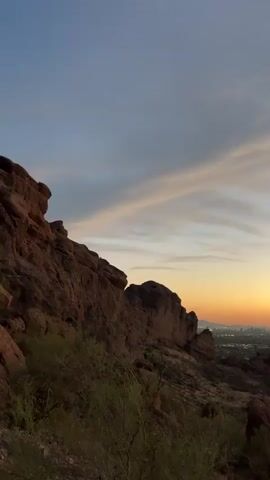 The video shows a time-lapse of a sunset over a city, as seen from a rocky, mountainous viewpoint. The sky transitions from a pale blue to shades of orange and red as the sun sets. The city lights begin to twinkle below. The foreground features rocky terrain with sparse vegetation.