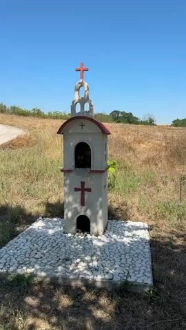 This media file captures a bright, sunny day in a rural setting, likely in Pravdino, Bulgaria. The scene opens with a close-up of a small, decorative bell tower, reminiscent of a chapel or shrine. It is painted white with a red roof and features red crosses. The base is covered in white pebbles. The camera then pans to reveal a dry, grassy field with scattered bushes and trees, suggesting a warm, possibly late summer or early autumn season. The sky is a clear, vibrant blue. The camera then moves to focus on a tree laden with small, round, reddish-orange fruits, possibly plums or cherries, hanging from its branches. The sunlight filters through the leaves, creating dappled shadows on the ground. The overall impression is one of natural beauty and tranquility. There is no visible text in the media file.