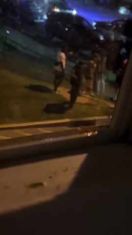 The video captures an outdoor scene at night, likely in Franklin Township, United States of America, viewed from an elevated perspective, possibly a window. A group of approximately five to seven individuals is gathered on a grassy patch that borders a paved area, which could be a sidewalk or a street.

In the background, several parked cars are visible, with their headlights and taillights illuminated, casting light across the scene. The people appear to be casually standing and interacting or conversing. One individual on the left side of the group is wearing a white t-shirt and dark shorts. Another person, positioned towards the center, is wearing a dark jacket with some light-colored text or a logo on its back that is mostly illegible, but seems to start with "80" or "BD". A distinct detail is one person on the right side of the group who is using crutches. The artificial lighting from the cars and possibly streetlights casts clear shadows of the people onto the grass. The foreground shows a portion of a window frame or ledge, indicating the recording location. The atmosphere appears calm and social, with no discernible strong emotions or urgent activity. The time of day is clearly nighttime, and there are no visible cues regarding weather conditions.
