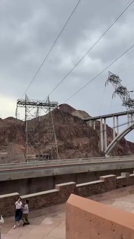 The video shows a view of the Hoover Dam and the surrounding rocky terrain. In the foreground, two people are standing and interacting. Behind them, a concrete walkway runs parallel to a large, imposing structure that appears to be part of the dam's infrastructure, possibly housing power lines or associated machinery. Large metal pylons and high-voltage power lines are visible, stretching across the landscape and connecting to the structures. In the background, a large arched bridge spans the canyon, with more pylons and power lines extending over it and disappearing into the rocky mountains. The sky is overcast with a thick layer of clouds, casting a diffused light over the scene. The overall impression is one of industrial scale and the interaction of human engineering with the natural environment.