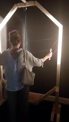 A woman with her hair in a bun, wearing a white blouse and a grey skirt, stands with her back to the camera. She is holding a wooden stick and interacting with a large, hexagonal wooden structure. The structure has multiple taut wires stretched across its frame, and the woman appears to be playing or experimenting with them. A light source within the structure illuminates the scene. To the left of the woman, a wall displays a colorful, distorted projection, possibly a result of light interacting with the wires or another element in the installation. The environment appears to be dimly lit, possibly an art exhibit or a science museum.