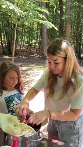 A woman and a young girl are standing outdoors in a wooded area. The woman, on the right, has long blonde hair and is wearing a white t-shirt and light blue denim shorts. She is taking a piece of food from a metal pot on a wooden table and placing it on a beige plate. The girl, on the left, has long dark hair and is wearing a white t-shirt with a blue Adidas logo. She is holding a beige plate with food on it and observing the woman. The metal pot contains what appears to be grilled meat and flatbread. The background shows trees and sunlight filtering through the leaves.