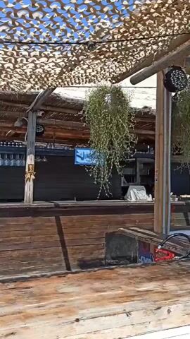A first-person perspective video shows a wooden structure with a thatched roof, possibly a beachside bar or restaurant. Hanging plants and some lighting fixtures are visible. Further along, there is a wooden walkway that leads to a cliffside overlooking the sea. A traditional fishing platform, known as a trabucco, is situated on the cliff. In the distance, people can be seen swimming and relaxing under an umbrella on a rocky shoreline. The sky is clear and blue.
