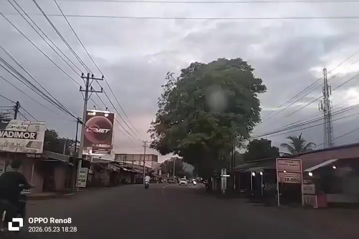 The video shows a road in a rural or suburban area. The camera is likely mounted in a vehicle, looking forward down the road. The sky is overcast, suggesting it might be early evening or a cloudy day. There are power lines running along the side of the road, and buildings on either side are a mix of small shops or houses with corrugated metal roofs. A large tree is on the right side of the road. There are a few vehicles and motorcycles moving along the road. The video seems to be taken in a region with some billboards, one visible for "Comet." The overall impression is of a simple road in a developing country, perhaps in Asia.