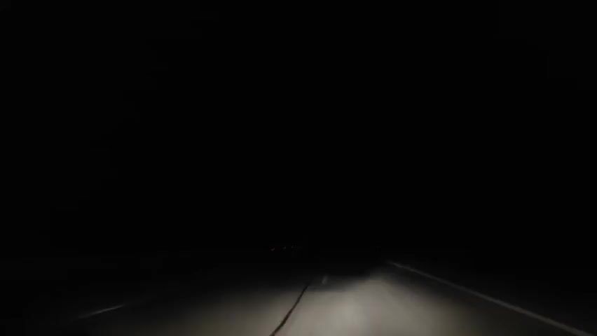 A driver's perspective from a vehicle on a dark highway at night. The vehicle's headlights illuminate the road ahead. Lightning flashes in the distance, revealing dark clouds and silhouettes of trees.