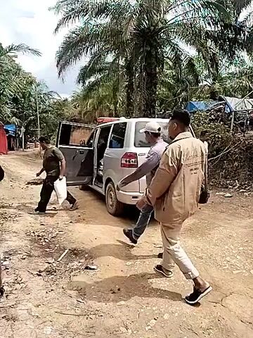 A medium shot shows the rear view of a white ambulance parked on a dirt road surrounded by palm trees and other vegetation. Four men are visible. One man exits the open side door of the ambulance, while another man in a tan jacket stands nearby with his back to the camera. A third man in a white shirt and dark pants stands to the right of the ambulance, looking at his phone. A fourth man walks away from the scene on the left side, carrying a white bag. The environment appears to be a rural or tropical area with sparse structures visible in the background. The dirt road is uneven with visible debris. The sky is overcast, casting soft lighting on the scene.