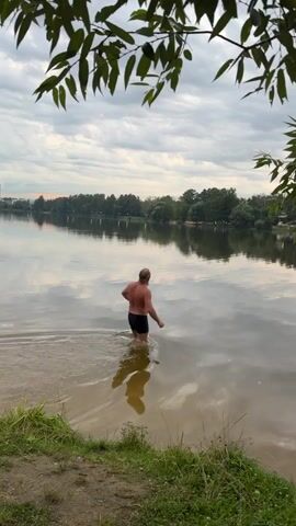 A man in black swim trunks walks into a lake. He is facing away from the camera, and his legs are submerged in the water up to his thighs. The water is calm and reflects the sky and trees. The shoreline is lined with grass and dirt. The sky is cloudy with some patches of blue. There are trees on the opposite shore.