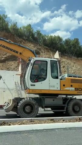 The video shows a highway construction site. An excavator is visible on the left side of the frame. A concrete wall is being constructed on a hillside. Formwork for the concrete wall is in place. A black car is in the foreground, and its reflection is visible on the windshield. Further down the road, a white truck passes by. In the background, there are trees and houses. The sky is blue with some clouds.