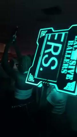 A person is holding a brightly lit sign that reads "Exclusive Ballers." The sign changes colors, alternating between green, blue, and purple. The person is in a crowded room with many other people. The room appears to be a party or event space with a ceiling fan and some decorations.