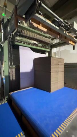 The video shows an industrial setting with a large, automated machine processing cardboard. The machine, painted in shades of green and brown, is positioned over a blue conveyor belt system. A substantial stack of flat, brown cardboard pieces sits on one of the blue conveyor belts. The machine features a series of rollers and a vertical chain-driven mechanism. To the left of the cardboard stack is a large, white, vertically oriented piece of material, possibly another form of cardboard or a backing. The camera angle is slightly low, looking up towards the machine. A green light is visible atop the machine. The background is a typical industrial warehouse setting with neutral-colored walls and ceiling, and metal meshing. The action involves the automated machine presumably preparing the cardboard for further processing or packaging.